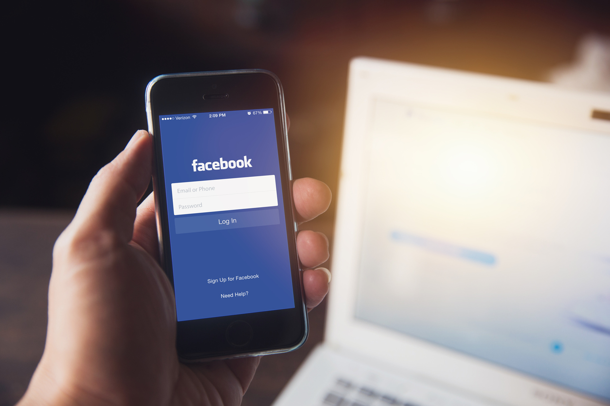 Facebook应用程序在iPhone与电脑笔记本电脑背景特写男性手握社交网络在智能设备的概念。